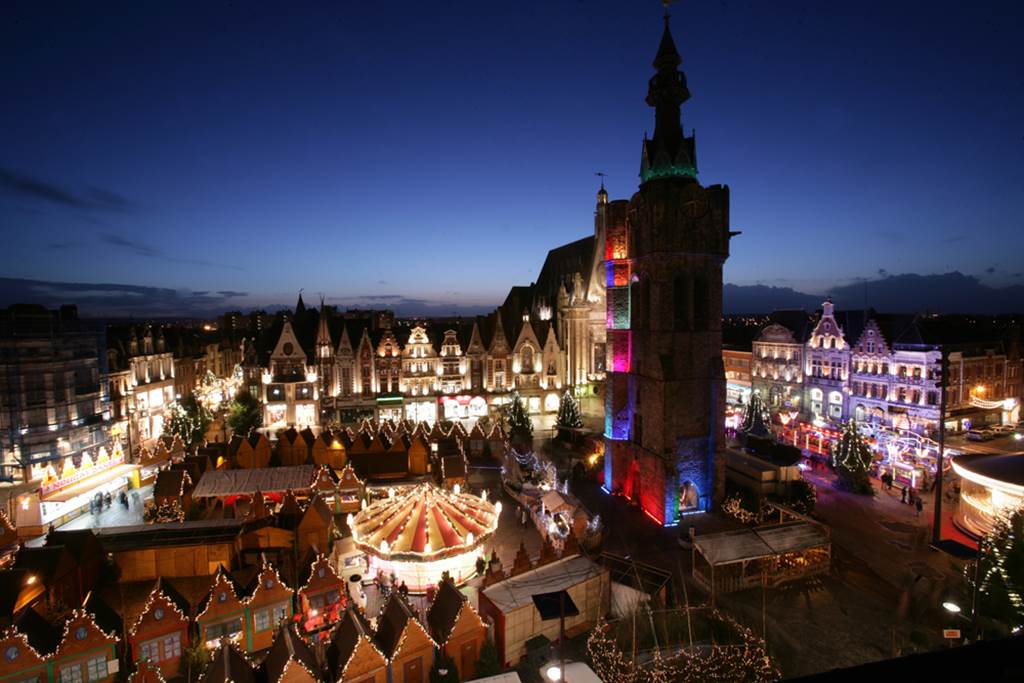 Aerial view of Bethune Christmas market with tall belfry above square with carousel and lit chalets and people 