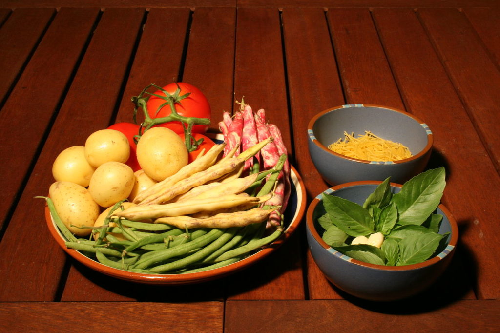 Pistou ingredients in three bowls with fresh vegetables in one, basil leaves and pine nuts in second and cheese in one behind on wooden slatted table