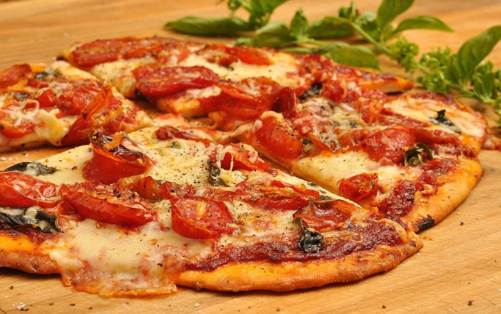 Pizza with tomatoes and cheese on table, cut up with green herb in background