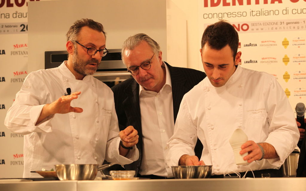 Alain Ducasse in jacket and shirt between 2 young chefs watching them as they prepare food