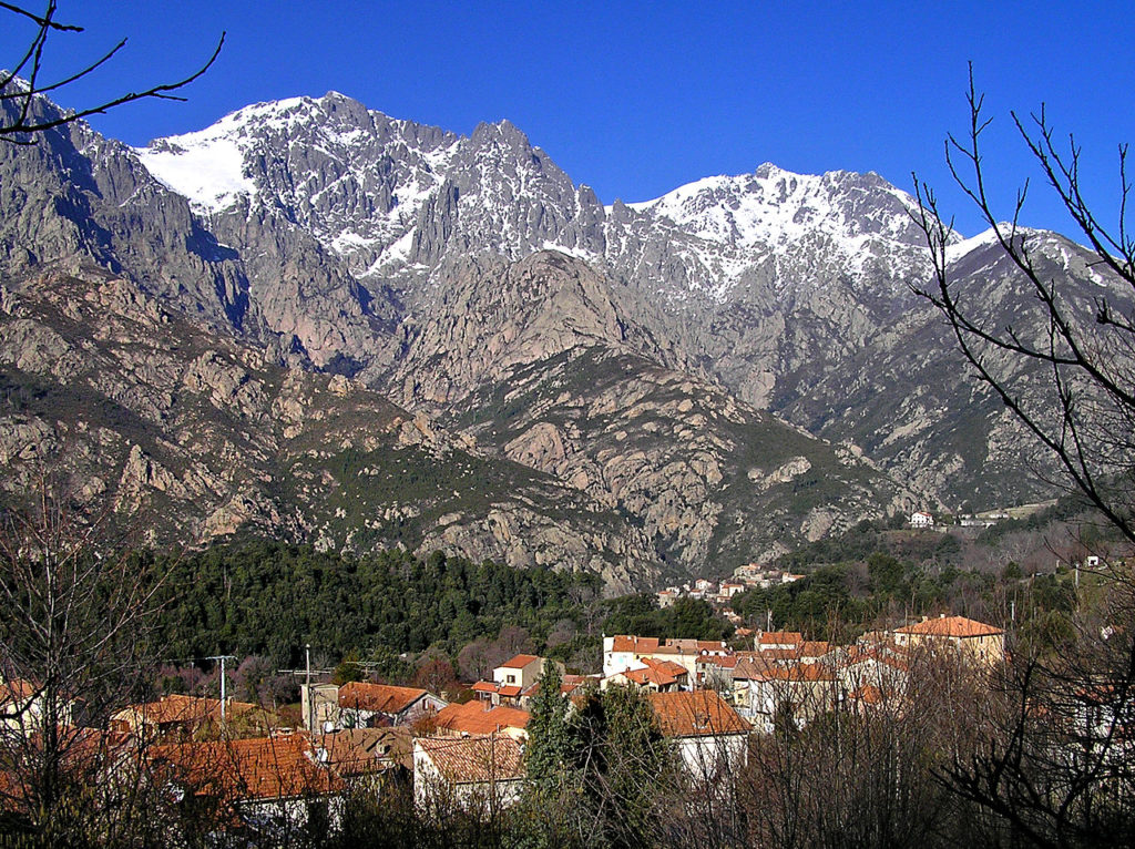 Bocognano, Corsica. Long view with snow covered mountains in the background and the small town in a valley below surrounded by trees. Red rooves and old houses