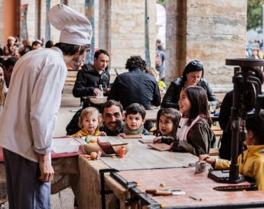Chef with huge toque in front of tables with kids behind at table height learning how to cook