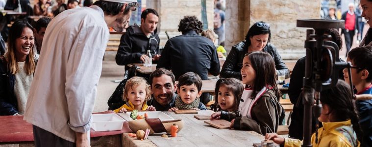 Chef with huge toque in front of tables with kids behind at table height learning how to cook