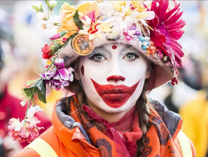 Full face of lady in Dunkik Carnival with multi coloured hat, costume from shoulder up and face painted white with huge grinning red mouth