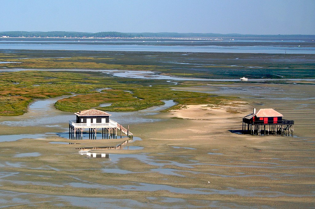 Bird watching cabins in Arcachon Bay on French Atlantic coast. Far veiw with two small 2-storey bird watching cabins in marshy lands with water and green patches, large water expanse and coastline in background