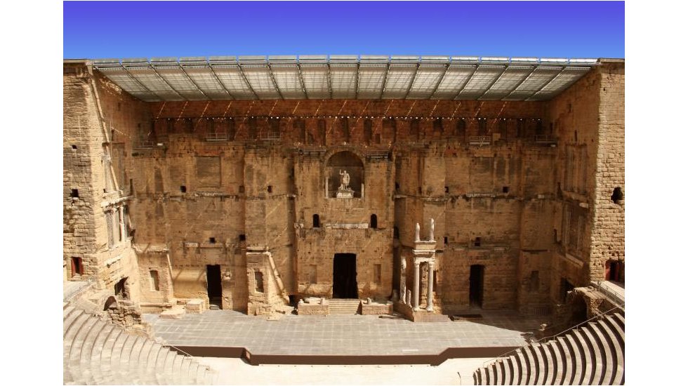 Full view of the stage at the Roman Theatre in Orange with its stage and stone backdrop with statues carved and freestanding. Steps onto stage