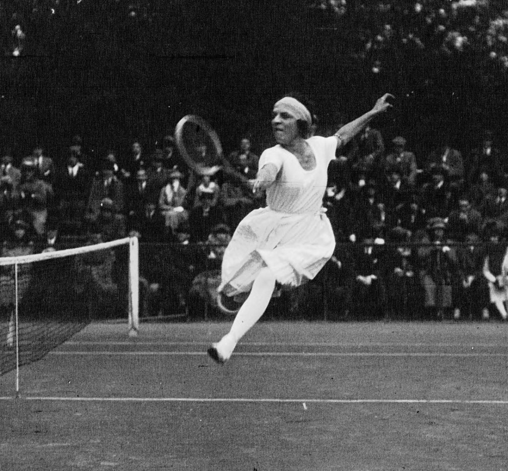 Suzanne Lenglen playing tennis in 1920. Old black and white photo of her in the air doing a backhand shot with one arm raised behind her. She's in long skirts