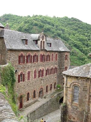 View of pilgrim hostel at Abbey Sainte Foy Conques. Tall 3-storey stone building with windows inthe slate roof by church with wooded hillsides behind
