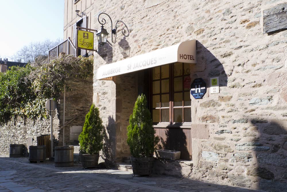 Stone wall and entrance of Auberge St jacques in Conques with logis sign, awning, and small trees flanking the entrance