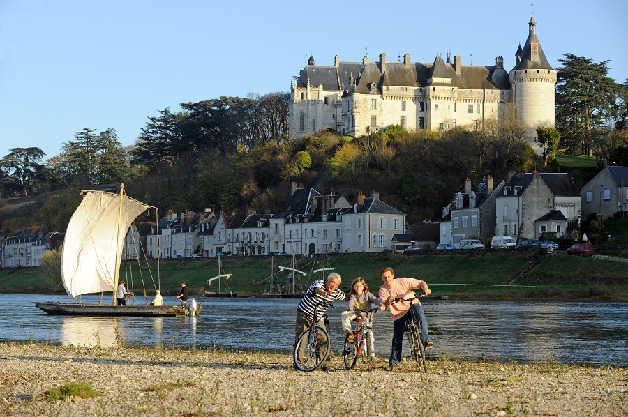 Three bicyclists pause by the river Loire with Chaumont perched on hill behind. They are facing towards the photographer and pointing out something to a child. Sailing boat of primitive kind on the river
