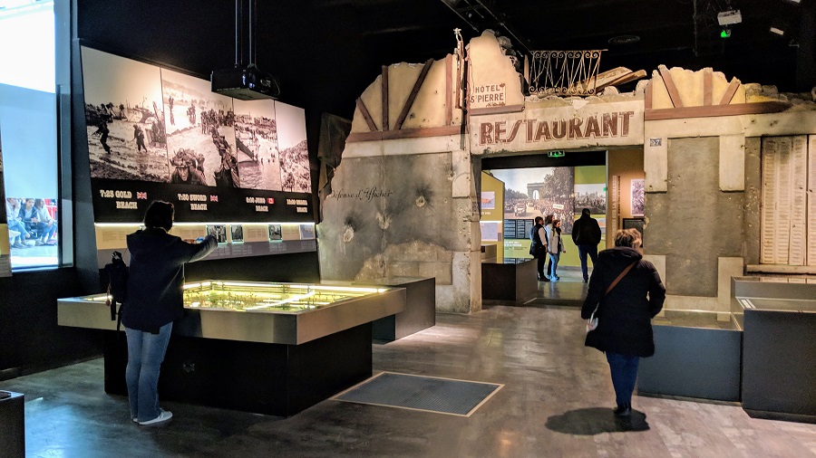 room setting at the Caen memorial with Restaurant written in large letters above entrance to second room at back, model in front and images around the walls Normandy Landing Beaches