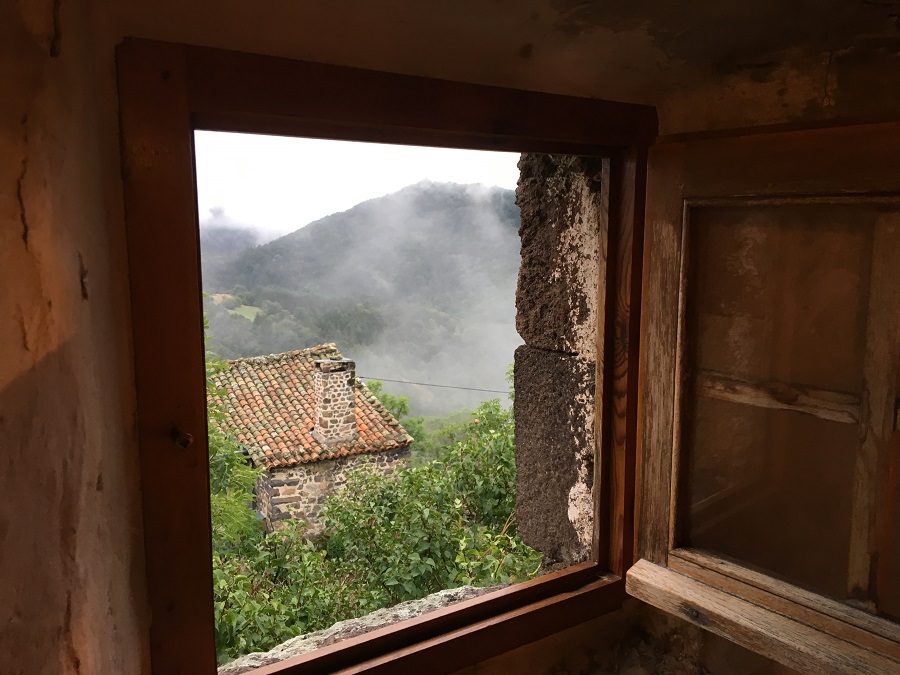 looking through small wooden window in stone walls at house with red roof and mist rising up
