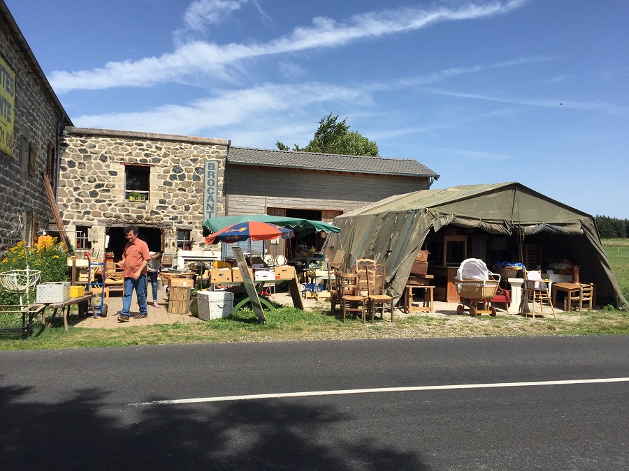 Brocante de Charreyrot in the Auvergne. Outside view from across the road with barn and large tent and old rustic furniture spilling out onto the grass and man walking off to left
