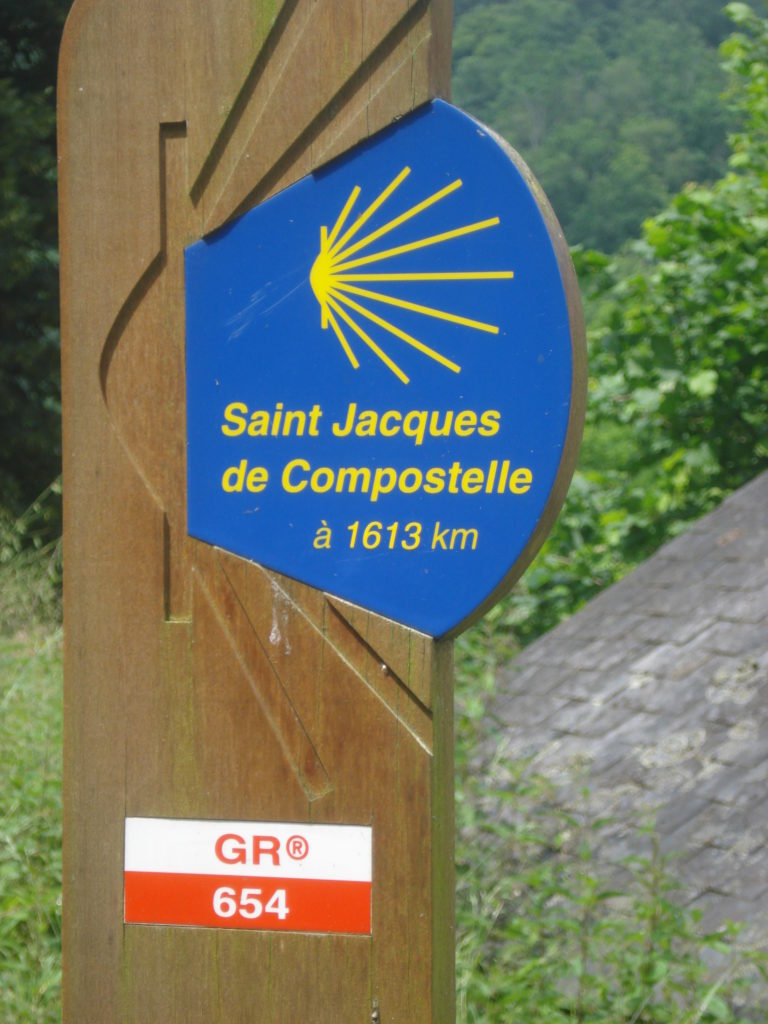 Public sign of St Jacques de Compostela. Blue background, idealised scallop shell on wooden post with path behind