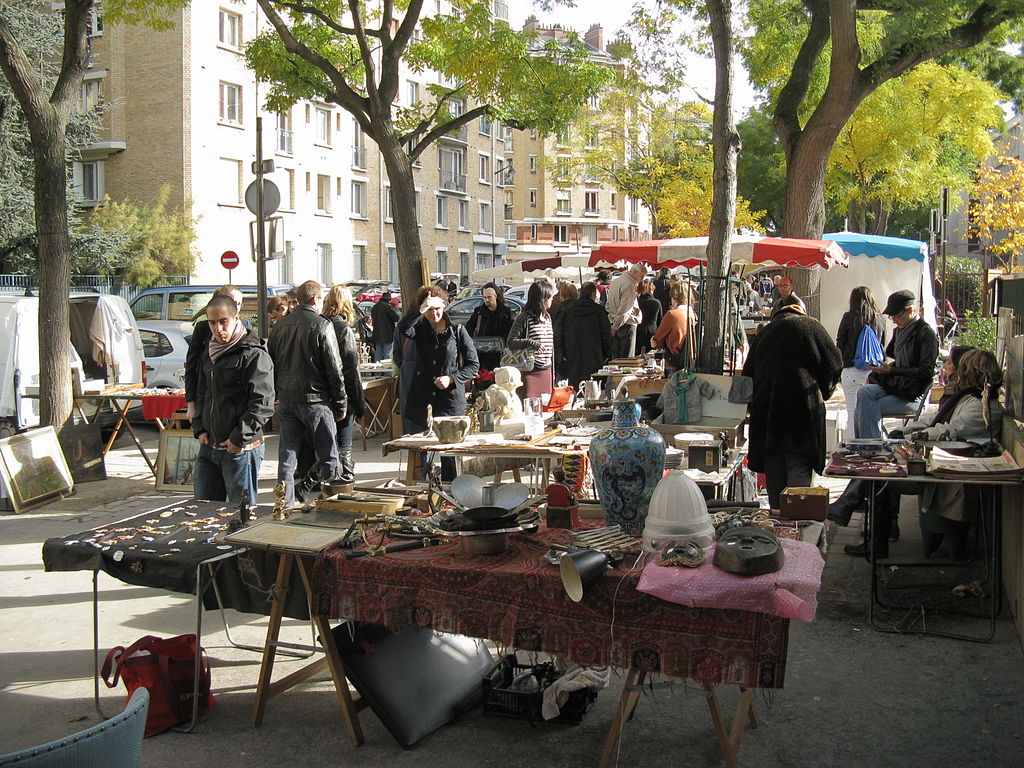 Puces de Vanves flea market Paris with people on street walking past tables set up on the pavement full of odd items from an African mask to badges