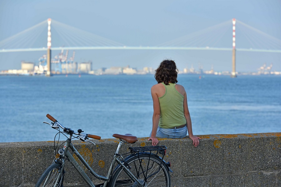 Back of girl sitting on a green bank with bcycle behind her looking out over the blue sea to the huge bridge at St Nazaire and the city beyond
