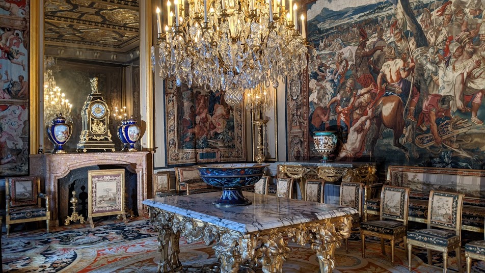 Queen Anne of Austria's Reception Room with huge chandelier lighting up ornate room with tapestries on walls, marble-topped table with gilt legs, fireplace with 3 clocks in blue lacquer on mantlepiece
