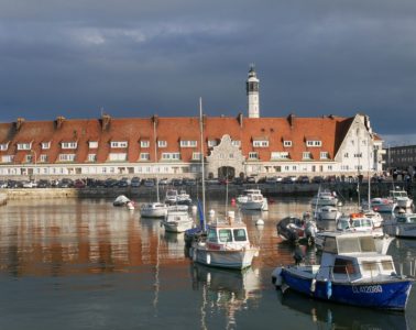 Calais harbour with boats bobbing in water and long building behind with red rooves and lighthouse in distance