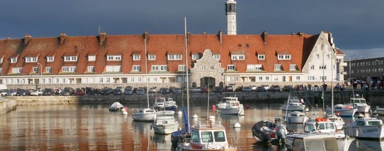 Calais harbour with boats bobbing in water and long building behind with red rooves and lighthouse in distance
