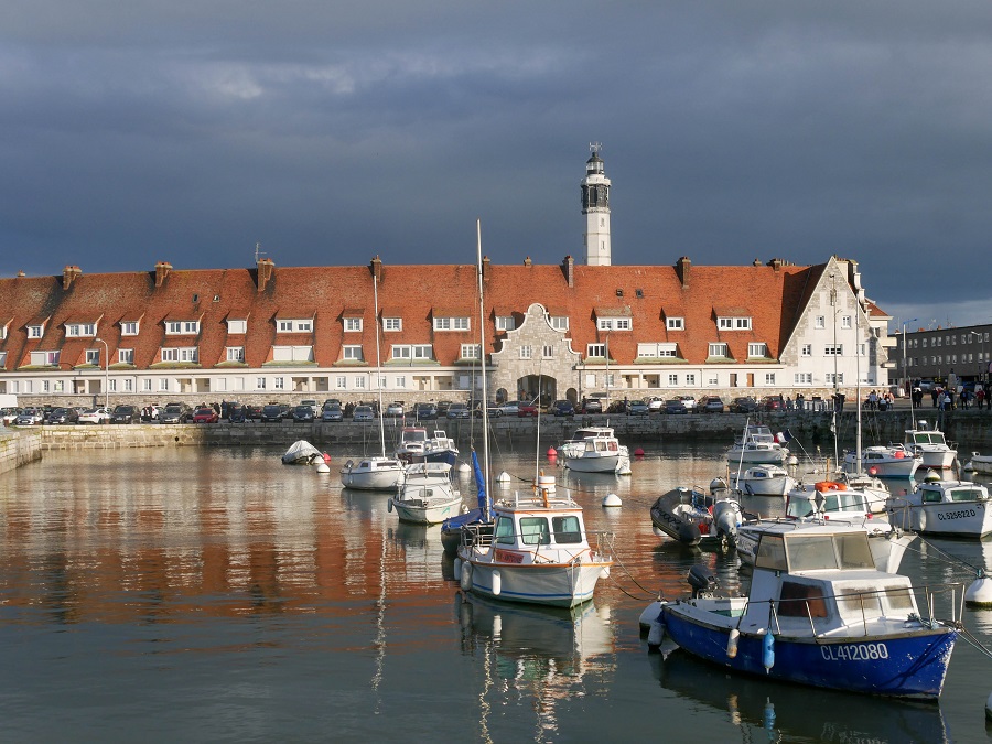 Calais harbour with boats bobbing in water and long building behind with red rooves and lighthouse in distance