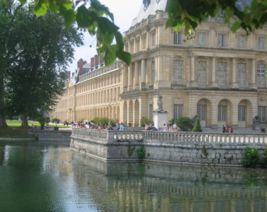 View of exterior of Fontainebleau Chateau. Corner view from lake with balustrade and people in front of mellow stone 4-storey building