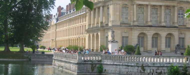 View of exterior of Fontainebleau Chateau. Corner view from lake with balustrade and people in front of mellow stone 4-storey building