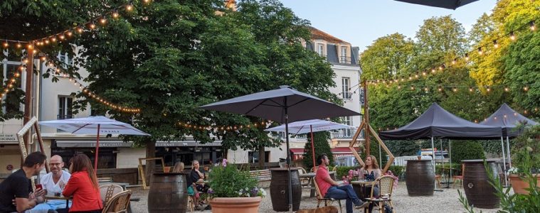 Courtyard bar in Fontainebleau with tables, chairs, parasols, people, a dog and barrels with trees and hotel behind