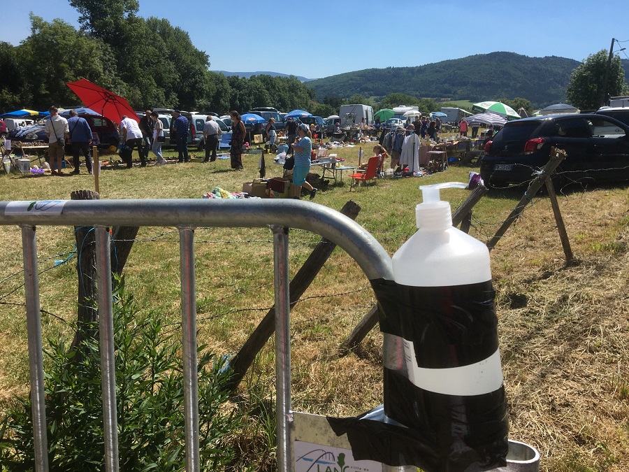 Plastic sanitising bottle taped to metal fence with stalls behind in a field at a vide grenier