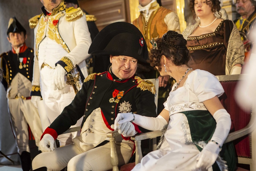Costumed actors playing Napoleon and Marie-Louise, holding hands in front of costumed courtiers