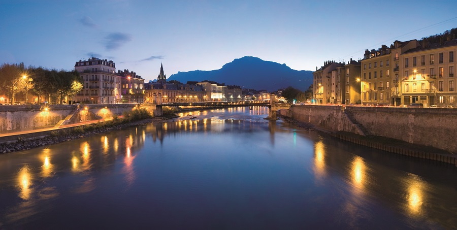 Grenoble at dusk from river in front with city on two banks reflected in the water and mountain behind