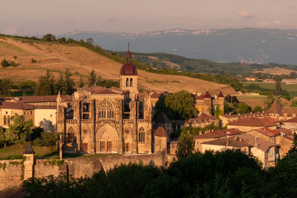 Saint-Antoine de l;Abbaye loking from afar to whole huge cathedral bathed in orange sunlight with hills and mountains behind