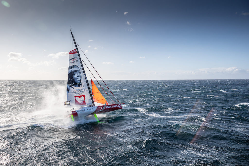 Sam Davies on her boat pre Vendee Globe 2020 showing boat going fast out to sea with spray at the back; sails covered in sponsors logos