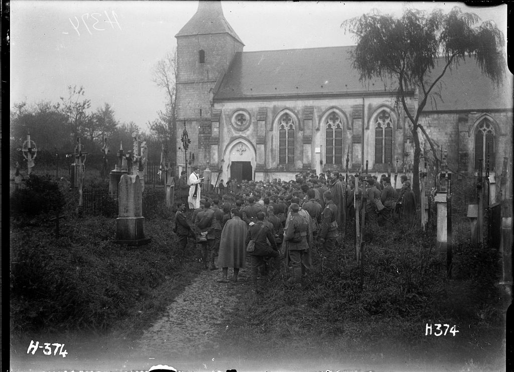 Black and white photo of All Souls Day 1917 in Pas de Calais with New Zealand soldiers gathered around a grave and a priest outside a church