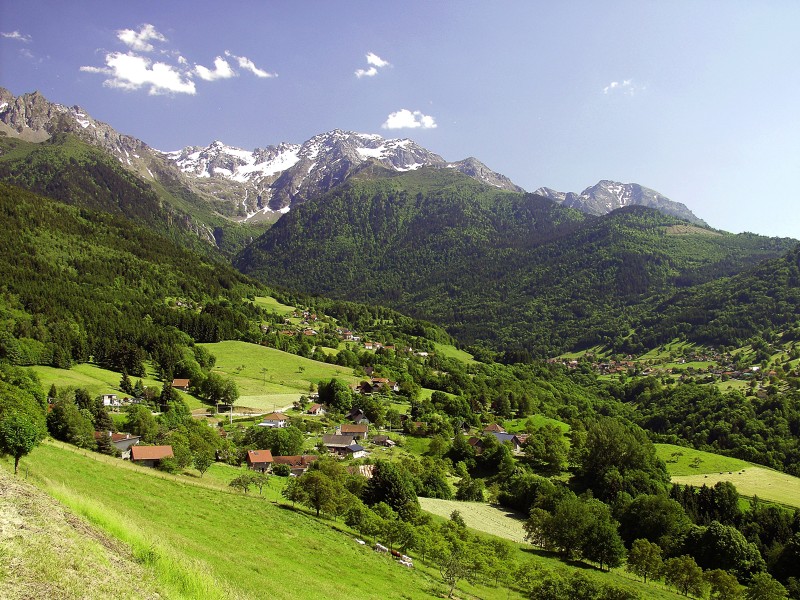 Balcons de Belledonne showing snow covered mountains in far background, green wooded hills middle and rolling sloping green pastures with red rooved farm buildings in front