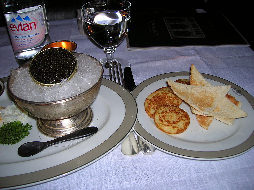Beluga caviar in small pot on ice in silver dish on plate with spoon at left. Blinis and sour cream on plate at right