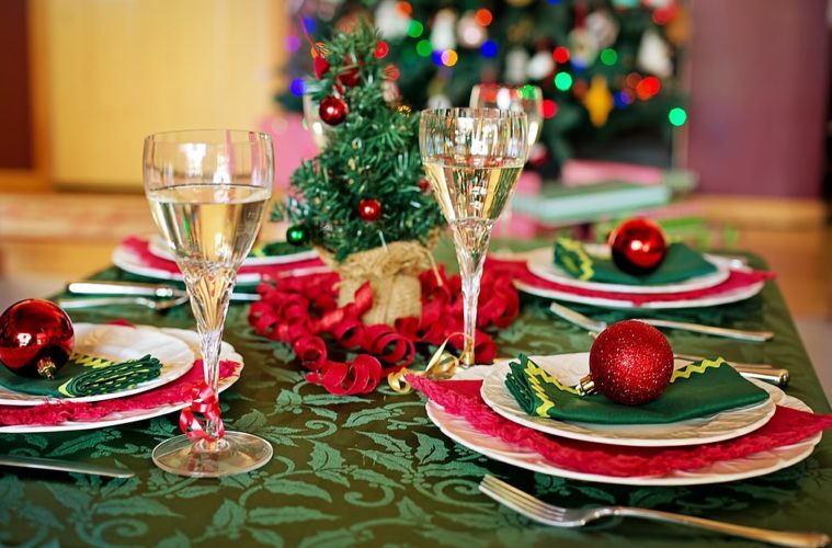 Tables laid for christmas with decorated tree in background, and table with green cloth, red baubles and ribbon and glasses of champagne