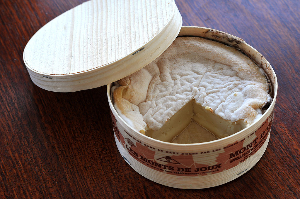 Round box of Vacherin Mont d'Or cheese with lide balanced on one side of the box and piece cut out to show wrinkly top and cheese inside