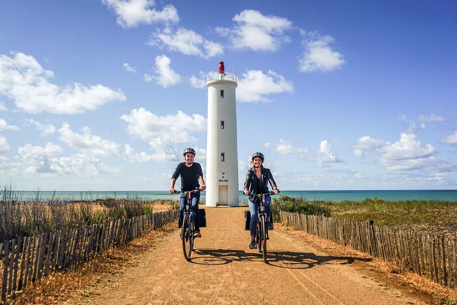 Two people, man and woman cycling towards camera on wide sandy path with lighthouse in distance