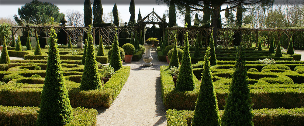 Gardens of William Christin showing gravelled paths between flower beds with box hedges and pyramid trees looking towards a little p;linth and gateway at back
