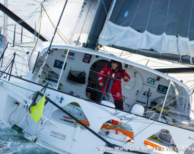 Jean Le Cam on his boat with pic from back showing him in main part of boat leaning sideways in sea spray