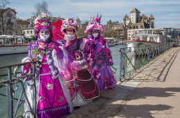 Annecy Venetian Carnaval showing four gorgeously dressed females in purple/pink costumes with headdresses and masks beside a canal