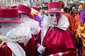 Limoux Carnival showing man in red costume with white ruff and red top hat withface painted in white with marks like a clows. He's surrounded by like-costumed people