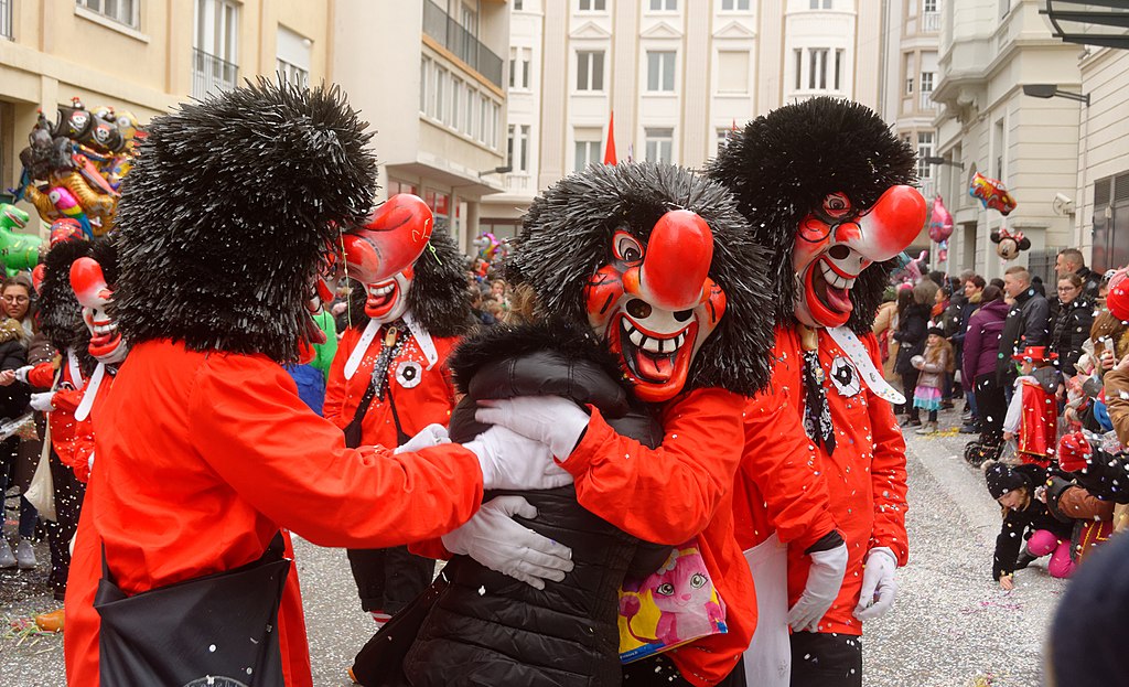 Mulhoue Carnival showing figures dressed in red with huge wigs embracing a passerby in the street