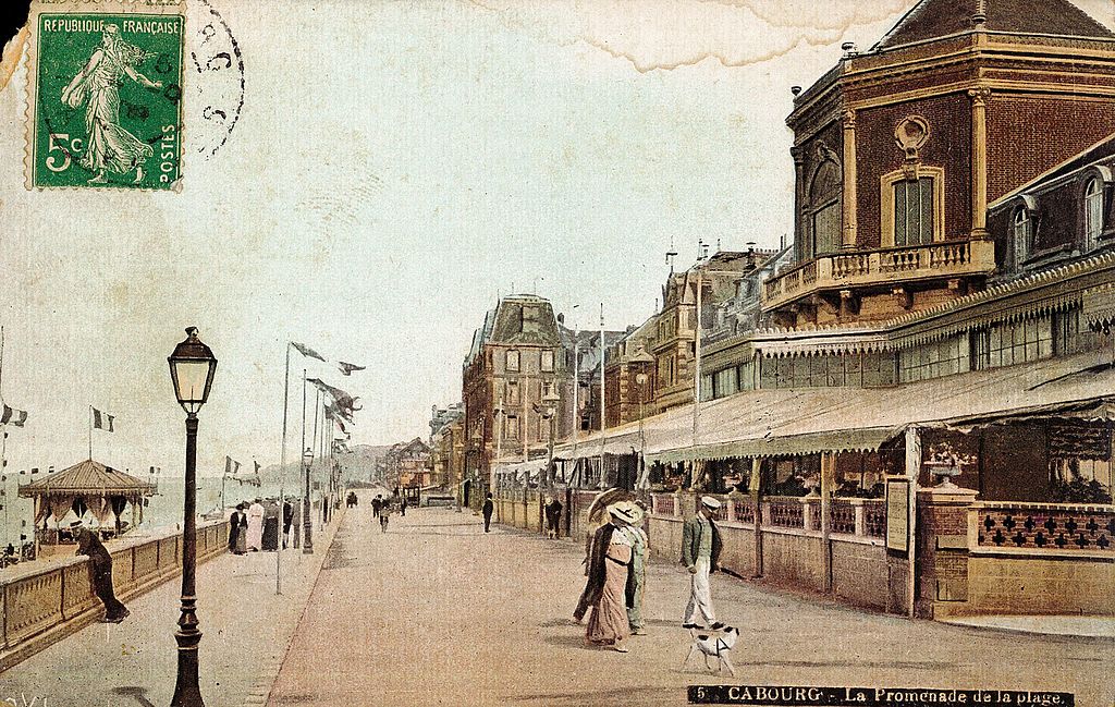Old hand coloured postcard of Cabourg in the 19th century showing sea promenade with gracious villas on right, and restaurant rterrace, wide promenade and ladies in long dresses walking