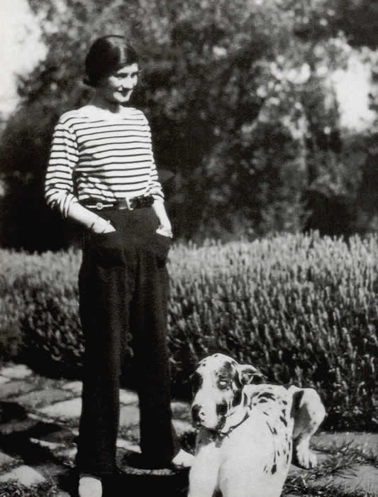 Balck and white photo of Gabrielle Chanel is marine style striped top and trousers in garden with dog