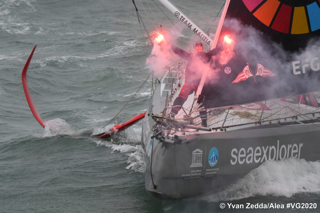 Boris Herrmann in boat finishing Vendee Globe just showing front of boat with Herrmann holding two red flares