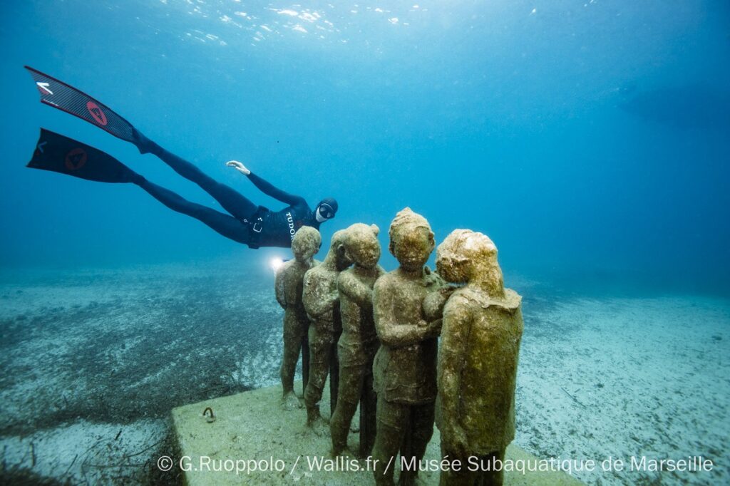 Diver in black behind group of sculptures of people on seabed