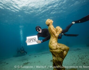 Diver in diving geat holding up sigh saying 'Open@ behind a sculpture planted onthe seabed