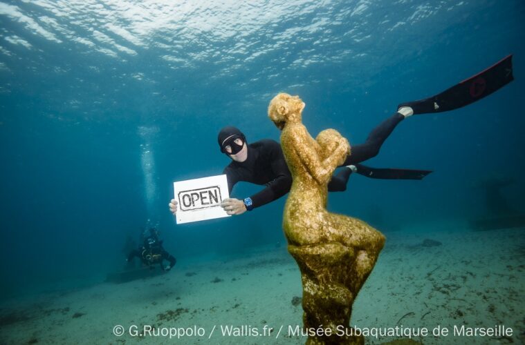 Diver in diving geat holding up sigh saying 'Open@ behind a sculpture planted onthe seabed