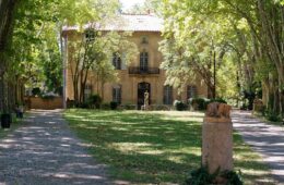 Mas du Jas de Bouffon of Cezanne in Aix-en-Provence. Warm stone 3-storey house with pitched red tile roof shaded by trees with green lawn in front and dappled view